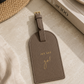 Katie Loxton Jet Set Go luggage tag in Taupe faux leather styled on a neutral tabletop with elegant travel-inspired accessories and warm natural lighting.