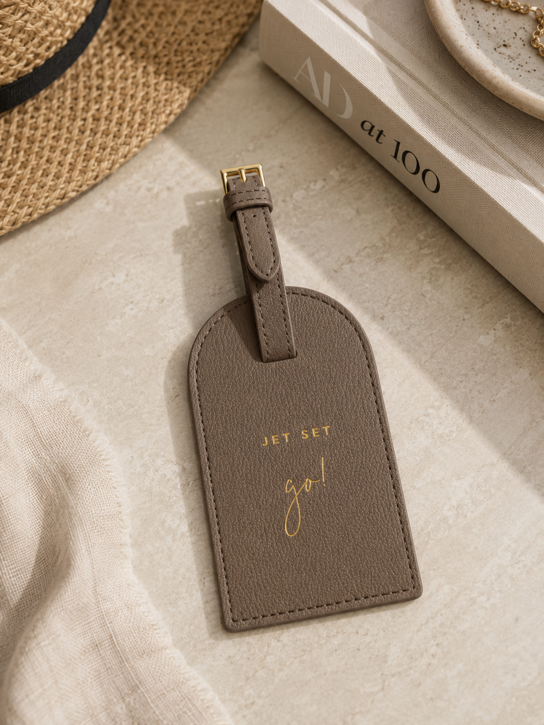 Katie Loxton Jet Set Go luggage tag in Taupe faux leather styled on a neutral tabletop with elegant travel-inspired accessories and warm natural lighting.