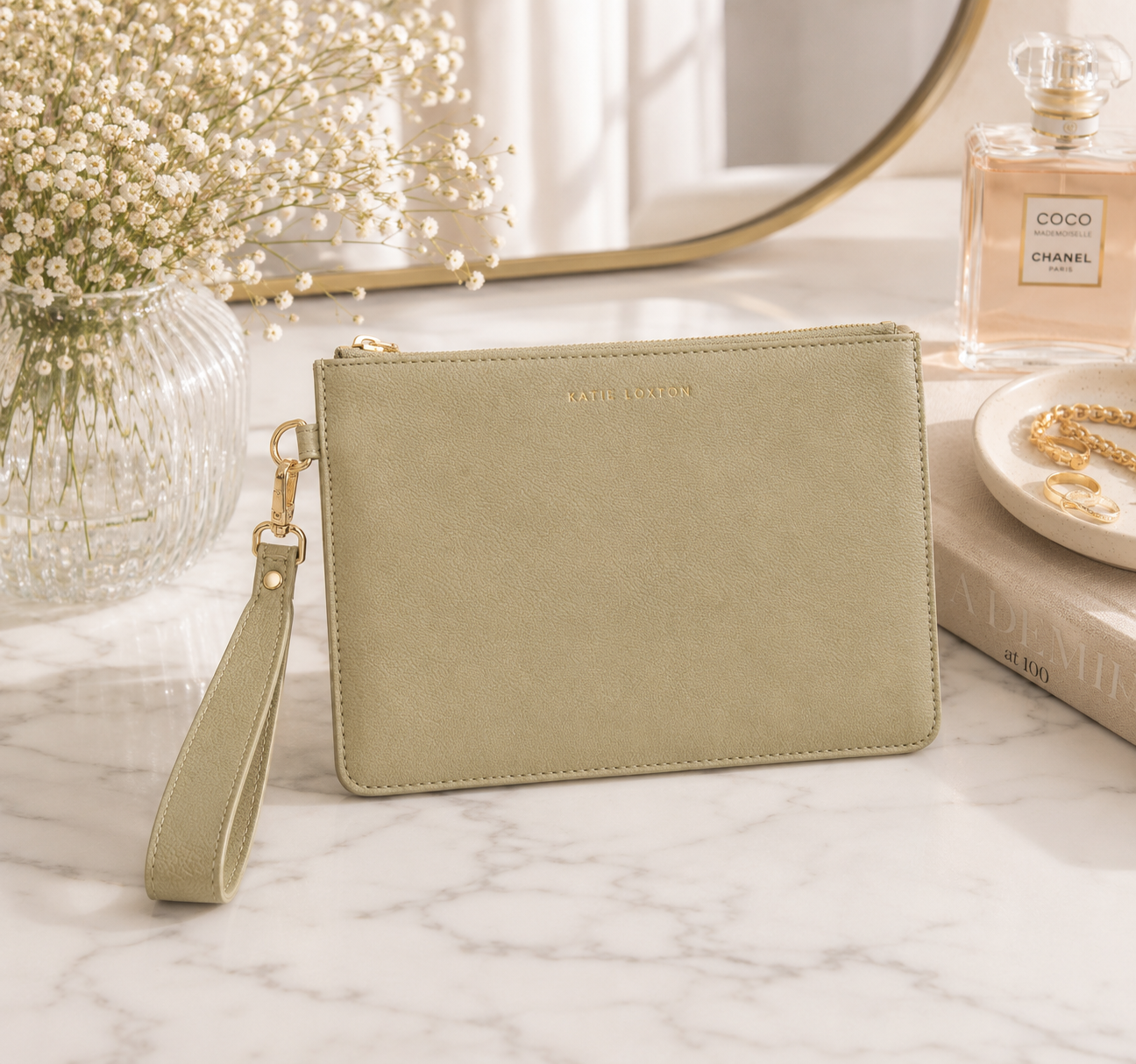 Katie Loxton Zana Wristlet Pouch in Sage Green faux leather styled on a luxury marble vanity with delicate gold jewelry, baby's breath, and elegant neutral décor.
