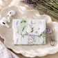 Lavender Meadows quilted AirPods case with delicate wildflower print and purple gingham lining styled with wireless earbuds on a marble tray.