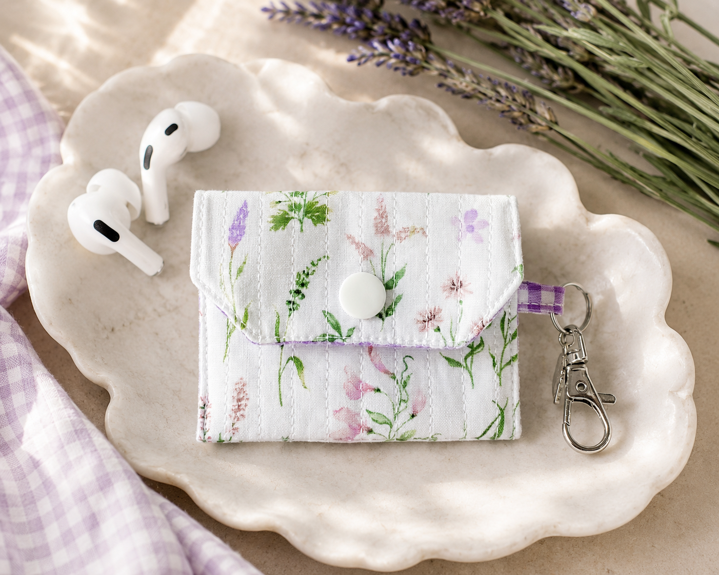 Lavender Meadows quilted AirPods case with delicate wildflower print and purple gingham lining styled with wireless earbuds on a marble tray.