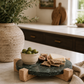 Round green marble serving board with a mango wood stand styled on a modern coastal kitchen countertop with artisan cheese, olives, crackers, and fresh figs.