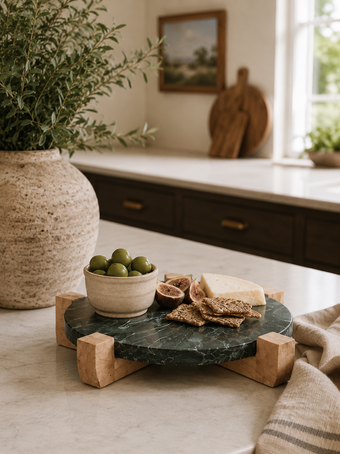 Round green marble serving board with a mango wood stand styled on a modern coastal kitchen countertop with artisan cheese, olives, crackers, and fresh figs.