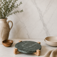 Round green marble serving board with a natural mango wood stand on a white marble background, showcasing a modern organic design for serving charcuterie, cheese, and appetizers.