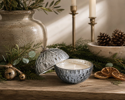 Mercury glass ornament candle with removable lid styled on a reclaimed wood table with fresh cedar and eucalyptus greenery, vintage brass bells, dried orange slices, pinecones, and warm neutral holiday décor in a designer-inspired Christmas setting.