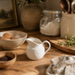 Rounded white ceramic creamer pitcher styled in a minimal editorial kitchen setting with layered neutral ceramics, soft linen textures, and warm natural light.