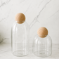 Tall and small modern clear glass pantry storage jar with sculptural natural cork ball lid on neutral studio background