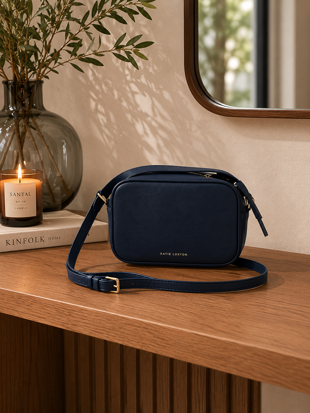 Navy Katie Loxton Isla Crossbody Bag styled on a warm oak console table in a quiet luxury interior with soft neutral decor and editorial lighting.