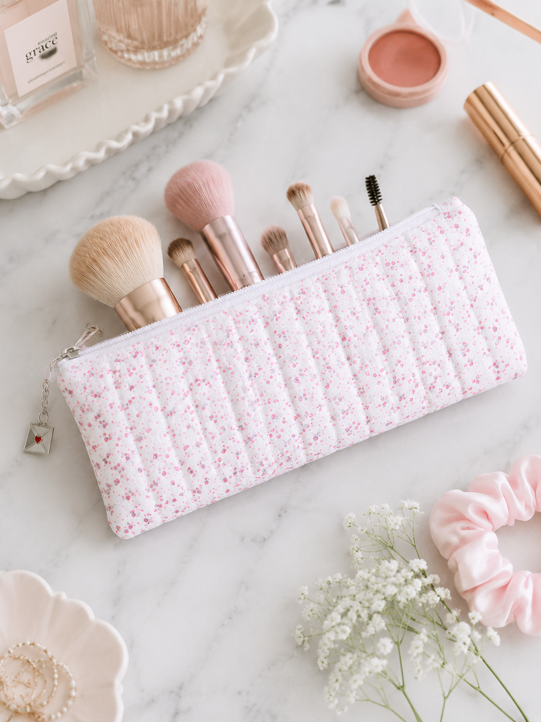 Pink floral quilted pencil case styled with makeup brushes and feminine beauty accessories on white marble countertop