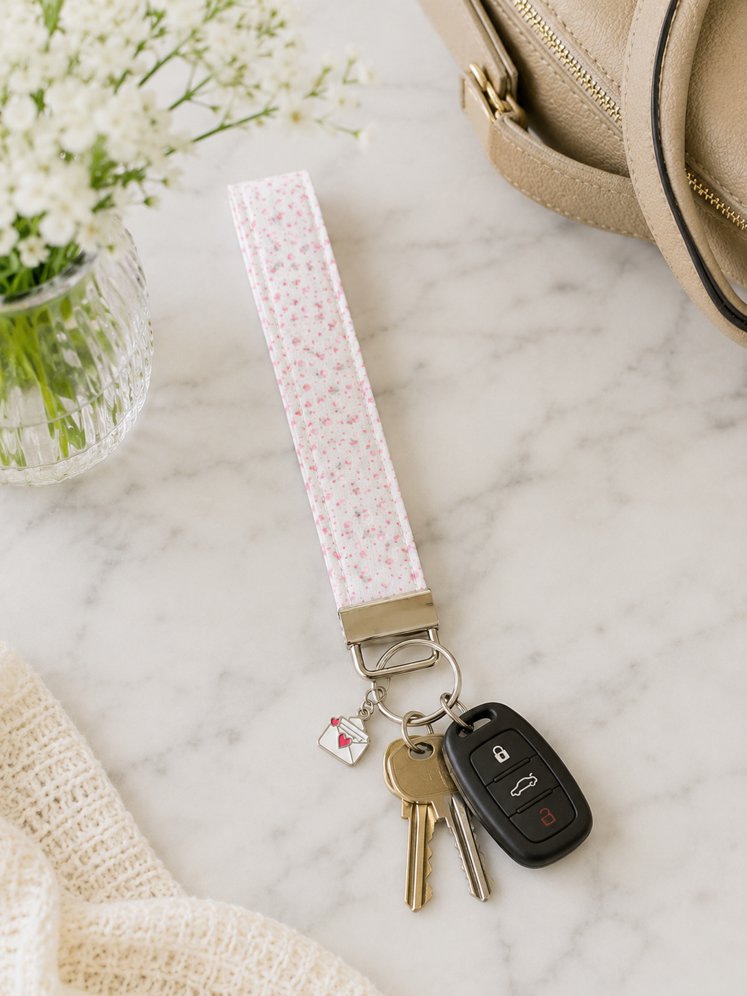 Pink floral wristlet keychain styled with car keys, neutral handbag, and soft feminine accessories on marble surface