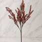 Rust brown faux astilbe stem displayed against a white marble studio backdrop, highlighting its natural texture and rich earthy color.
