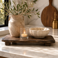 Dark rustic wood riser board styled with a textured ceramic vase, candle, and sculptural bowl on creamy marble kitchen countertops.