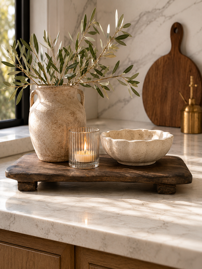 Dark rustic wood riser board styled with a textured ceramic vase, candle, and sculptural bowl on creamy marble kitchen countertops.