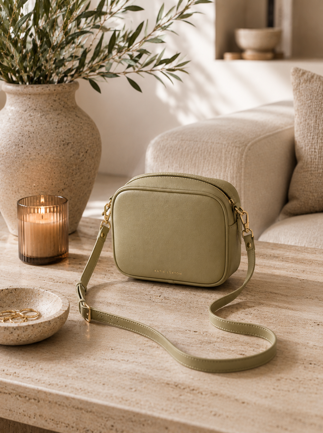Sage green Katie Loxton Isla Crossbody Bag styled on a travertine coffee table with olive branches, candlelight, and warm neutral California-modern living room decor.
