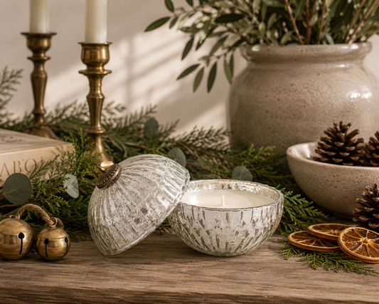Silver mercury glass ornament candle with removable lid styled on a reclaimed wood table with fresh cedar and eucalyptus greenery, vintage brass bells, dried orange slices, pinecones, and warm neutral holiday décor in a designer-inspired Christmas setting.
