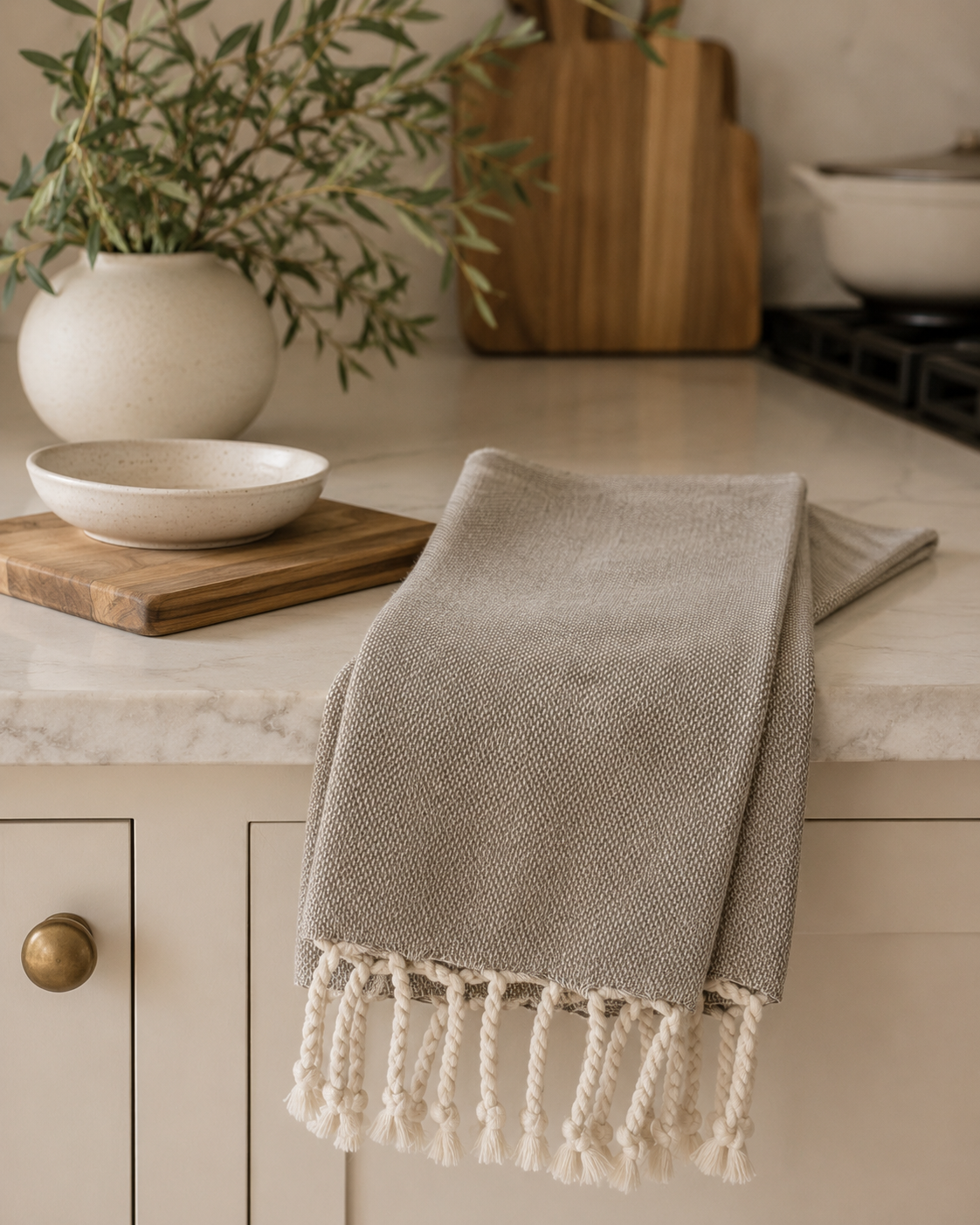 Solid taupe woven Haman tea towel with tassel fringe draped over a marble kitchen countertop in a modern California-inspired kitchen.