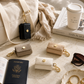 Neutral Katie Loxton AirPod cases styled in a luxury travel flat lay with a cream canvas tote, passport, coffee cup, sunglasses, gold jewelry, and marble surface in warm natural light.