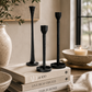 Three black cast iron taper candle holders styled on stacked neutral coffee table books with warm candlelight, olive branches, ceramic decor, and soft linen accents in a cozy luxury interior.