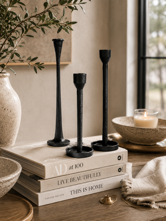 Three black cast iron taper candle holders styled on stacked neutral coffee table books with warm candlelight, olive branches, ceramic decor, and soft linen accents in a cozy luxury interior.