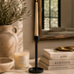 Black cast iron taper candle holder styled on a neutral console table with a cream taper candle, stacked coffee table books, artisan ceramics, olive branches, and warm natural light in a cozy modern interior.