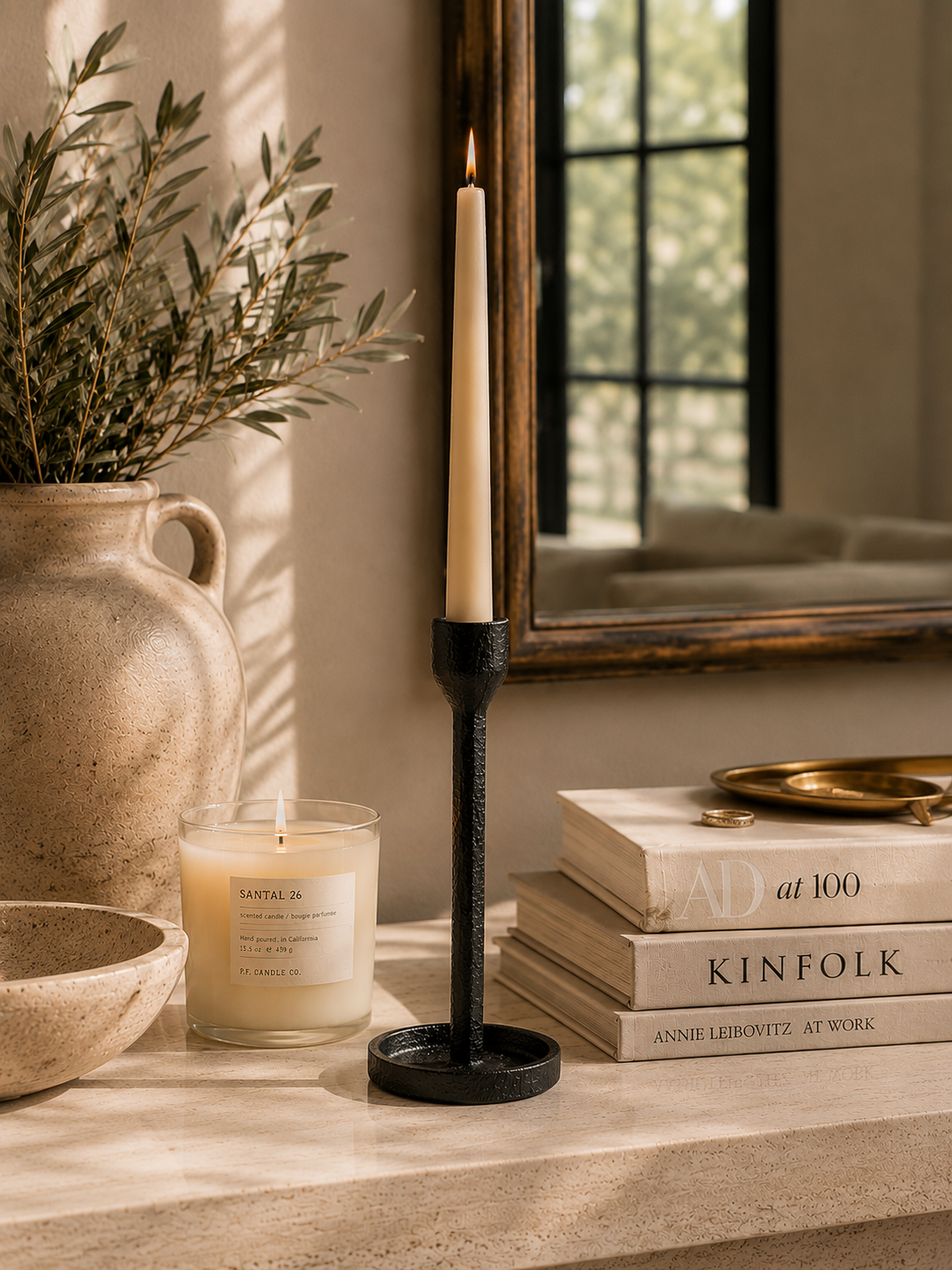 Black cast iron taper candle holder styled on a neutral console table with a cream taper candle, stacked coffee table books, artisan ceramics, olive branches, and warm natural light in a cozy modern interior.