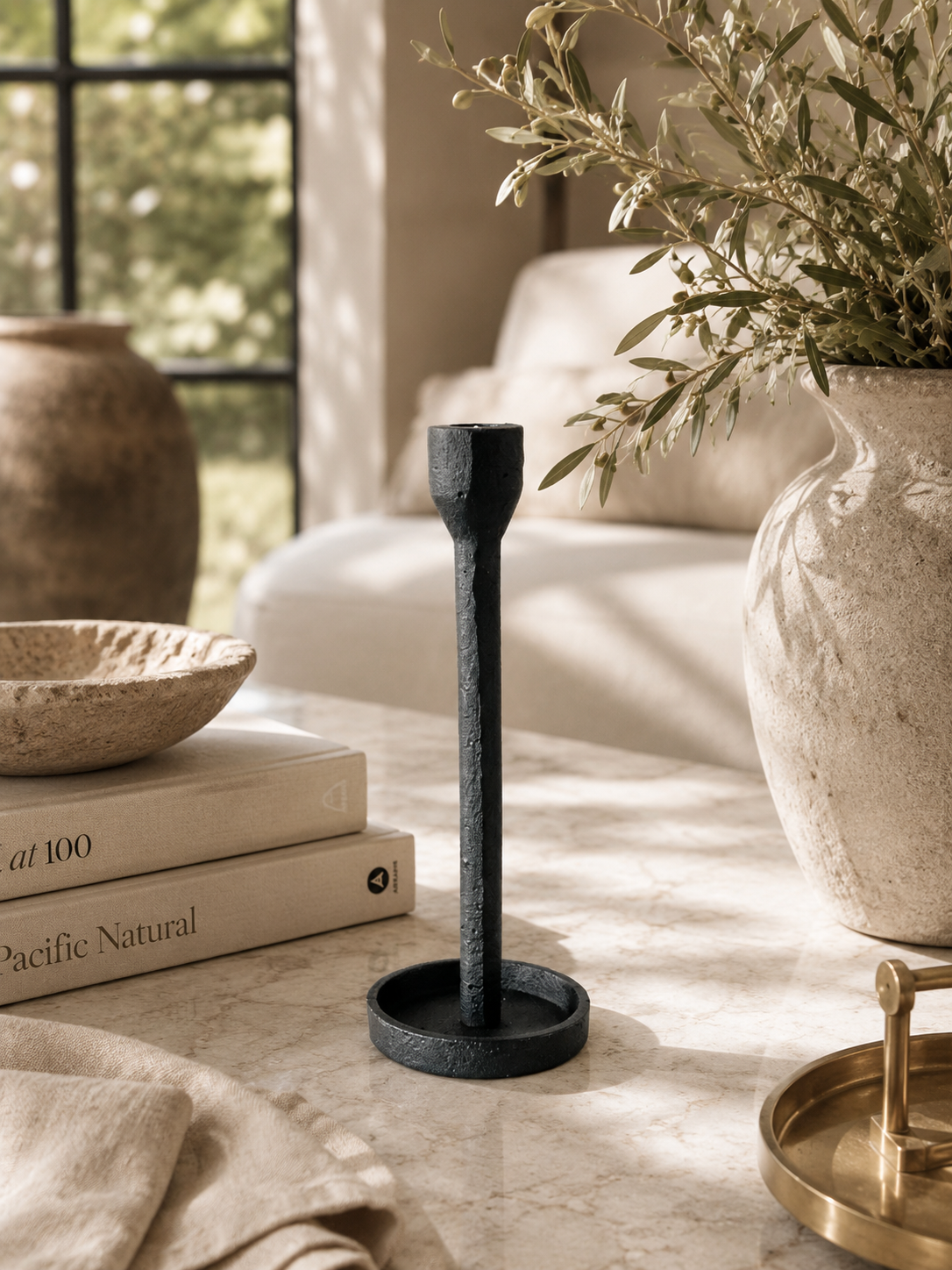 Black cast iron taper candle holder styled on a neutral coffee table with stacked design books, textured ceramics, olive branches, and soft natural light in a warm modern living space.