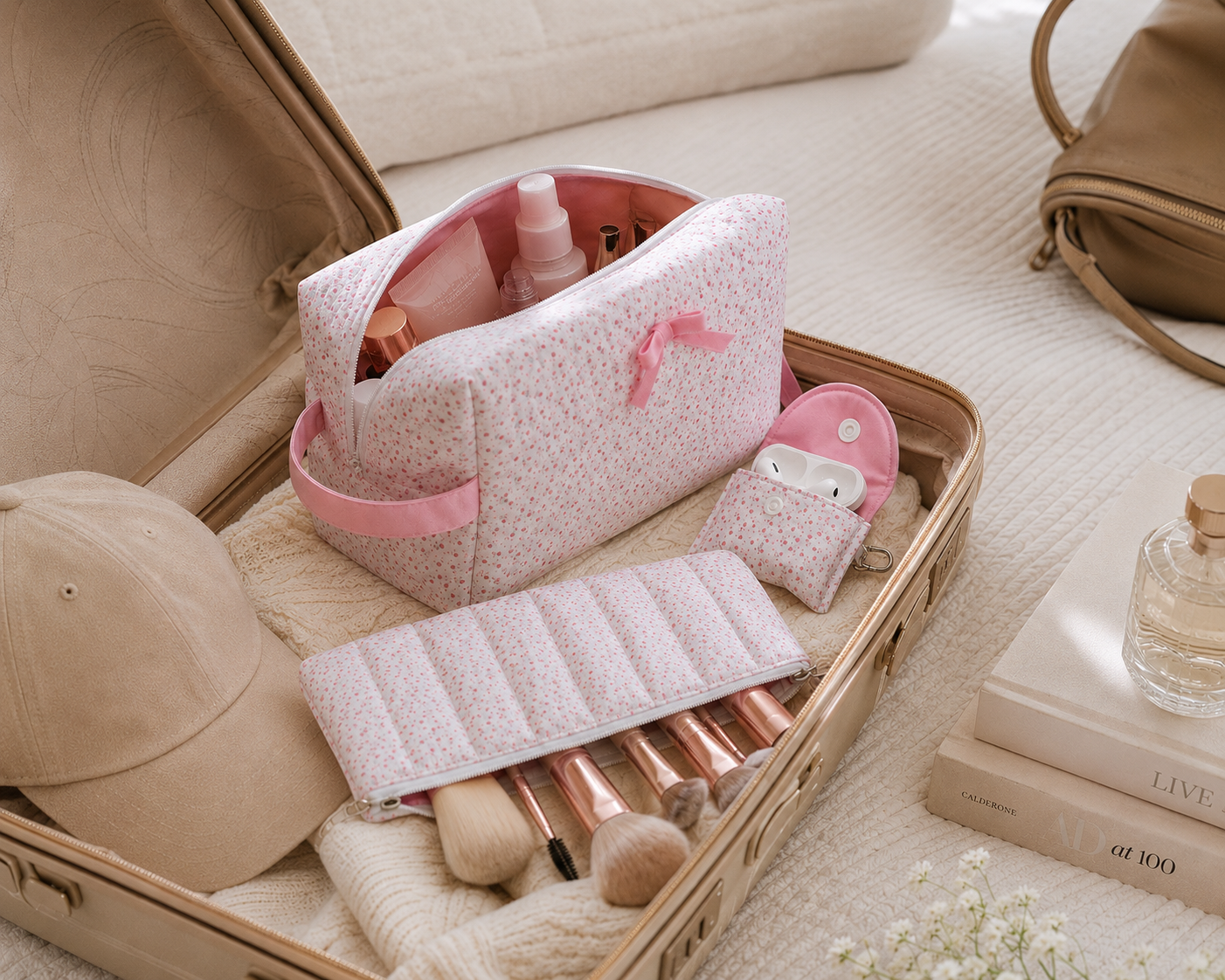 luxury travel packing scene featuring pink floral cosmetic bags, matching AirPods pouch, and makeup brush case styled inside an open neutral suitcase with soft feminine travel essentials and coffee table books.