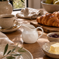 Small white ceramic creamer pitcher styled on a marble countertop beside croissants, espresso cups, soft linen, and olive branches in a warm California-modern breakfast kitchen.