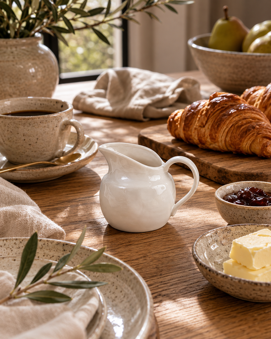 Small white ceramic creamer pitcher styled on a marble countertop beside croissants, espresso cups, soft linen, and olive branches in a warm California-modern breakfast kitchen.