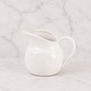 Minimal white ceramic creamer pitcher photographed on a clean marble surface with matching marble backdrop in a soft neutral studio setting.