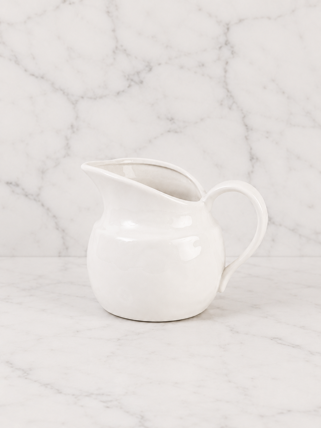 Minimal white ceramic creamer pitcher photographed on a clean marble surface with matching marble backdrop in a soft neutral studio setting.