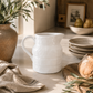 White stoneware pitcher styled in a warm California modern kitchen with olive branches, artisan bread, linen textiles, and creamy marble countertops.