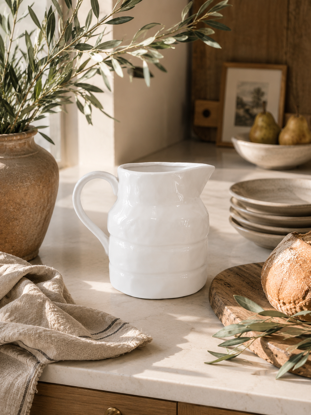 White stoneware pitcher styled in a warm California modern kitchen with olive branches, artisan bread, linen textiles, and creamy marble countertops.