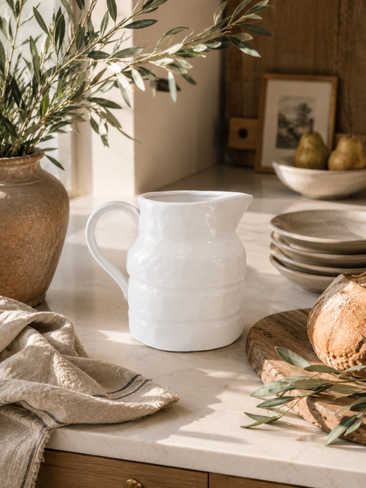 White stoneware pitcher styled in a warm California modern kitchen with olive branches, artisan bread, linen textiles, and creamy marble countertops.