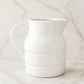 White stoneware pitcher with a glossy ceramic finish styled on a creamy marble surface with soft natural light and a minimalist neutral backdrop.