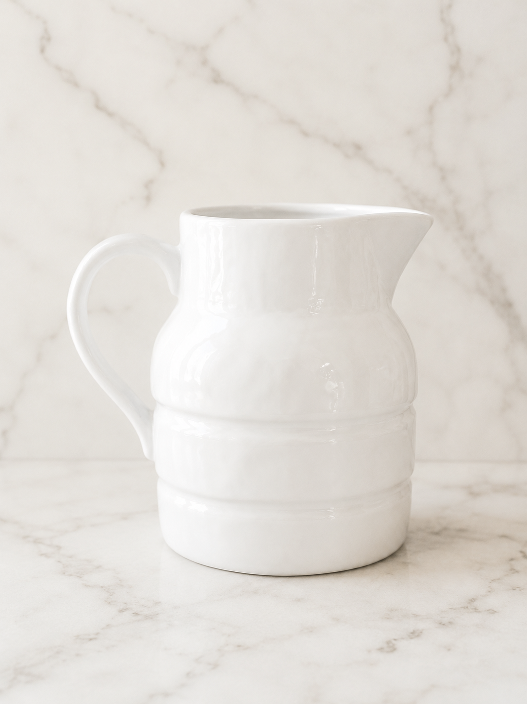White stoneware pitcher with a glossy ceramic finish styled on a creamy marble surface with soft natural light and a minimalist neutral backdrop.