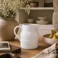 White stoneware pitcher styled on a rustic wooden table beneath warm oak shelving with fresh pears, artisan ceramics, linen textiles, and soft California modern natural light.