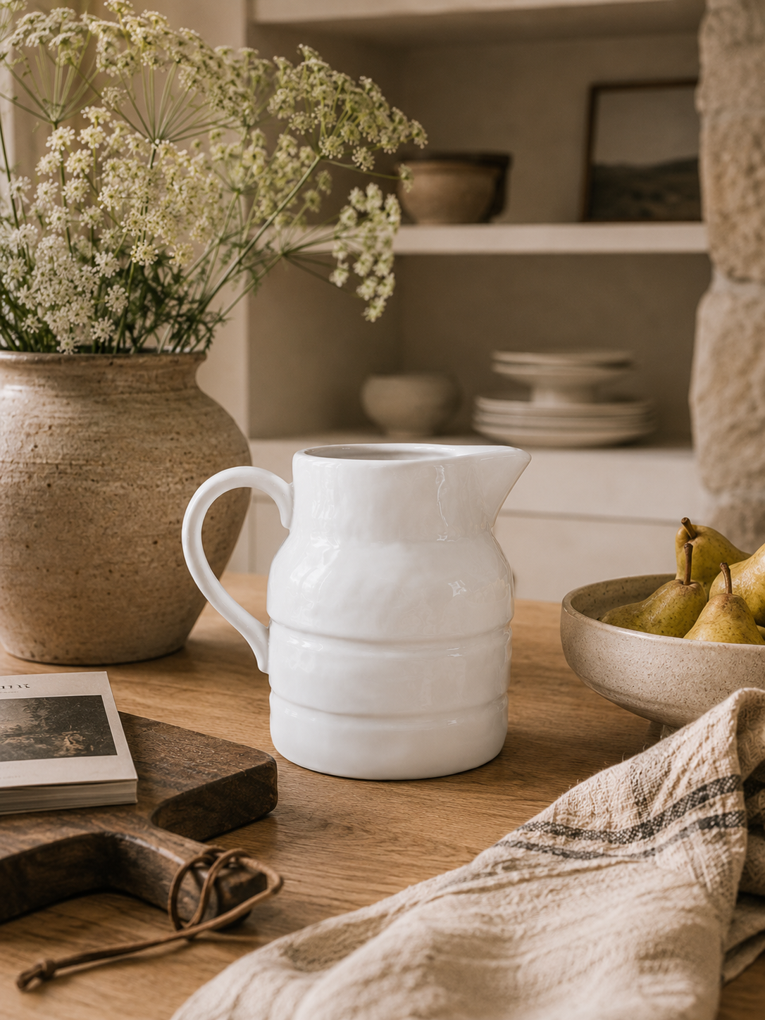 White stoneware pitcher styled on a rustic wooden table beneath warm oak shelving with fresh pears, artisan ceramics, linen textiles, and soft California modern natural light.