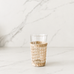Handwoven rattan wrapped drinking glass styled in a minimal marble studio setting with soft shadows and airy neutral tones.