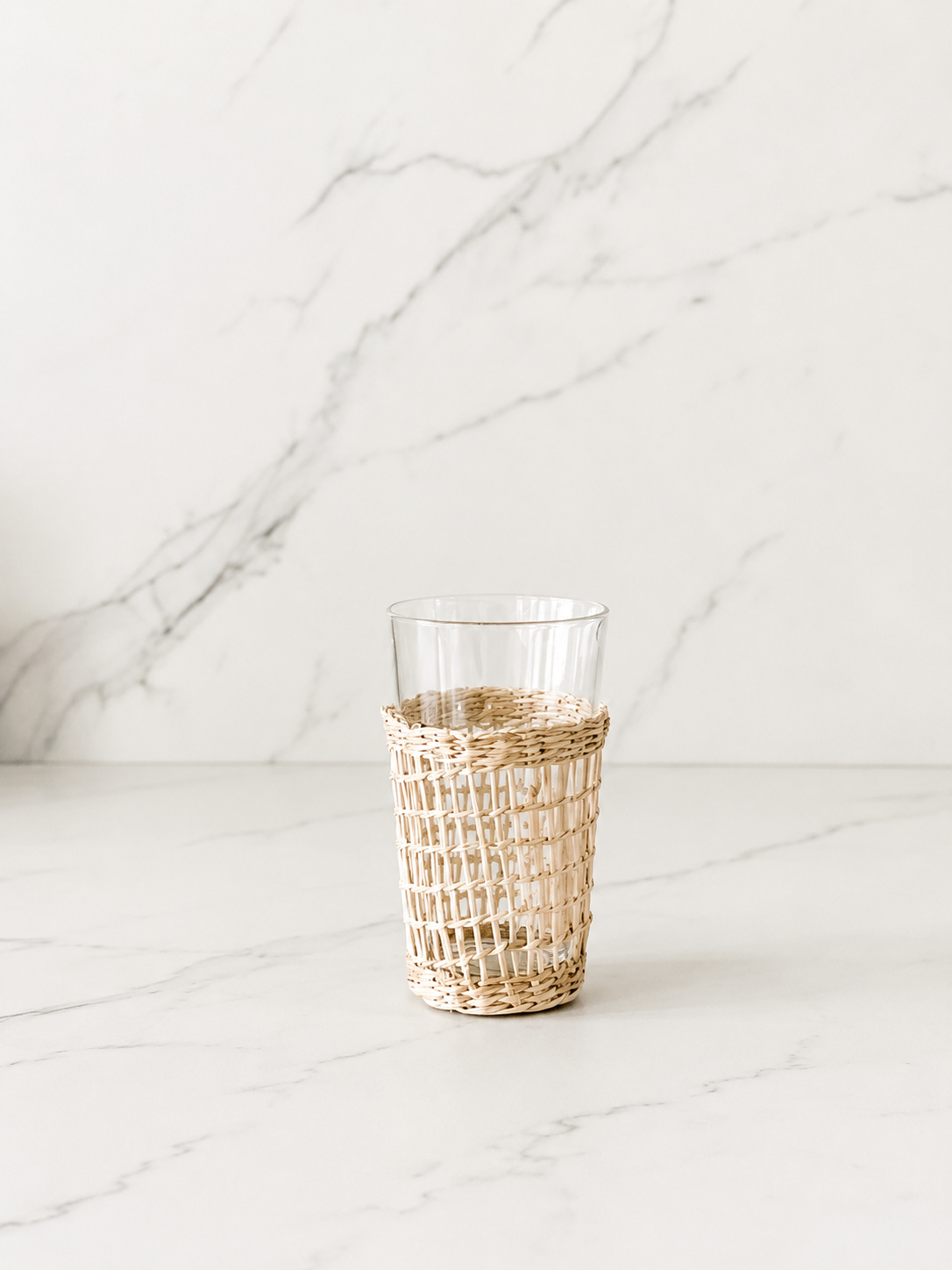 Handwoven rattan wrapped drinking glass styled in a minimal marble studio setting with soft shadows and airy neutral tones.