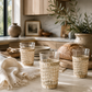 Three woven rattan drinking glasses styled on a creamy marble countertop in a warm California modern kitchen with artisan bread, linen textiles, olive branches, and soft natural sunlight.