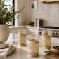 Three woven rattan drinking glasses styled on a creamy marble kitchen island in a warm California modern kitchen with olive branches, stacked ceramic dishes, linen textiles, and soft natural sunlight.