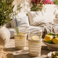 Two woven rattan drinking glasses styled on a sunlit outdoor patio table with lemons, olives, linen textiles, and Mediterranean-inspired decor in a warm California coastal setting.