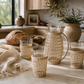 Woven rattan glass pitcher styled with matching woven drinking glasses on a creamy marble countertop in a warm California modern kitchen with artisan bread, olive branches, and soft natural sunlight.
