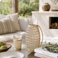 Glass pitcher with woven rattan sleeve styled on creamy marble countertops beside artisan bread, olive branches, and neutral California modern kitchen decor.