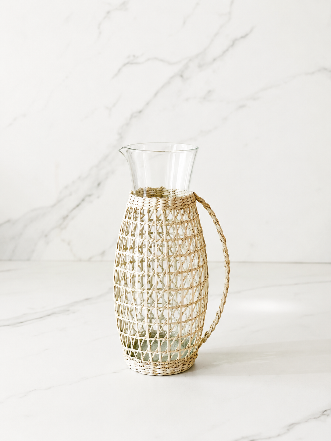 Handwoven rattan wrapped glass pitcher styled in a minimal marble studio setting with soft shadows and airy neutral tones.