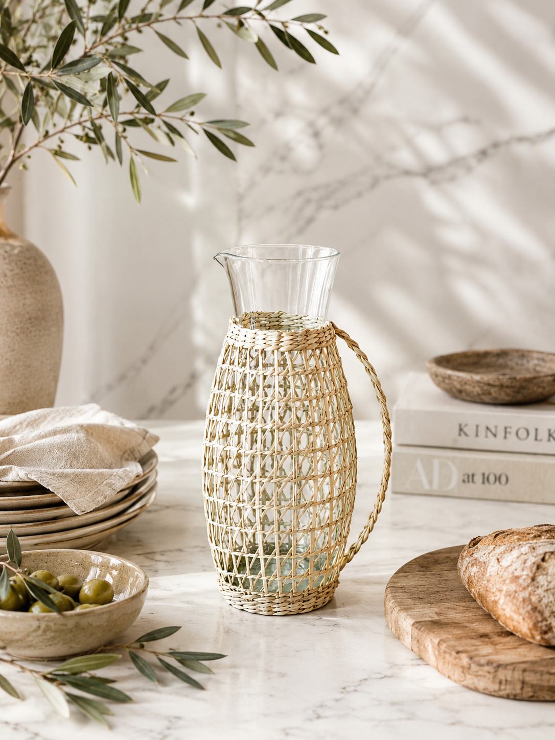 Clear glass pitcher with handcrafted woven rattan sleeve photographed on a soft marble backdrop with clean studio styling and natural light.